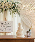 Good Vibes Neon Sign LED Wall Decor With Warm White Glow For Living Room And Weddings Boho Minimalist Modern Farmhouse Backdrops USB Powered 3D Art Gift 16.1x8.3 Inch displayed at wedding reception with floral arrangement and cake.