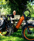 Blue Fat Tire Electric Bike For Epic Mountain Trails And Smooth City Cruising With Killer Torque Hella Durable Frame Long Range Battery Plus Utility Bike Bag For Zero Hassle Daily Rides parked on grass next to a man working on a laptop.