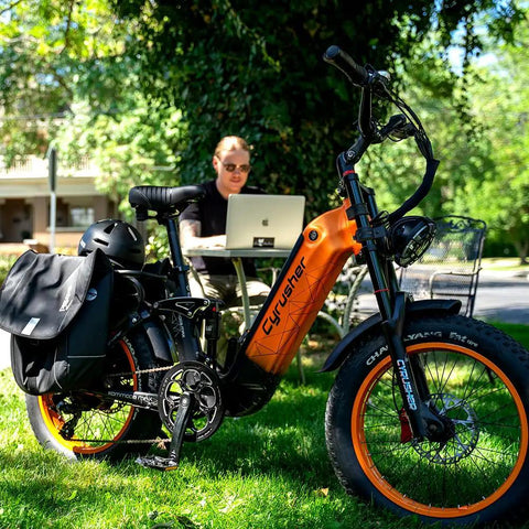 Blue Fat Tire Electric Bike For Epic Mountain Trails And Smooth City Cruising With Killer Torque Hella Durable Frame Long Range Battery Plus Utility Bike Bag For Zero Hassle Daily Rides parked on grass next to a man working on a laptop.