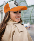 Woman wearing orange and white trucker cap with "NO CRYING ON THE YACHT" text, smiling in urban setting