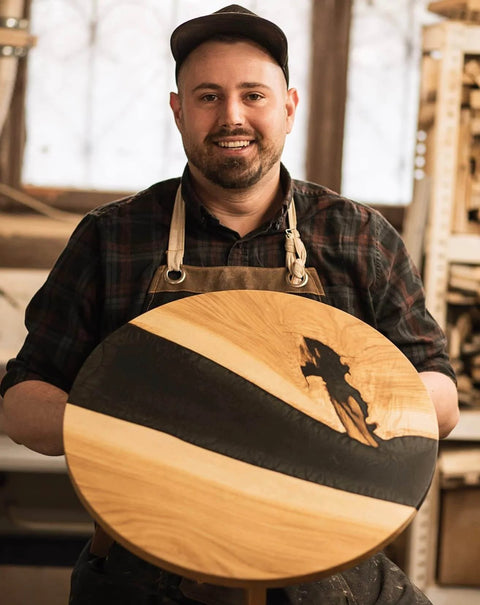 Woodworker holding a round tabletop with black epoxy inlay, showcasing woodworking craftsmanship.
