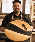 Woodworker holding round wooden table with black resin inlay in workshop, showing craftsmanship and design.