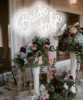 Bride To Be Neon LED Sign White Letters Light For Bridal Shower And Bachelorette Party Wall Decor Reusable Neon Wall Art Gift For Engagement Celebrations Party Ready 17.7 x 12.2 Cm above floral arrangement at bridal event