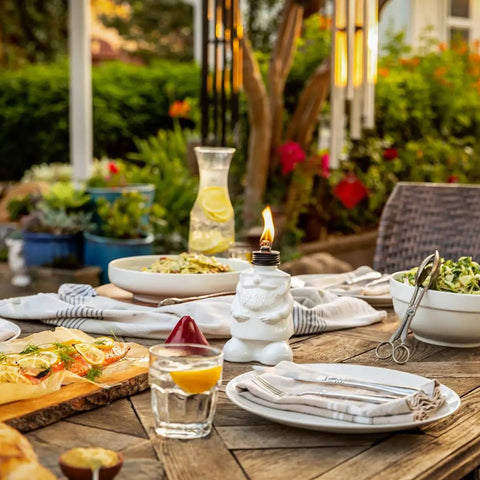 White gnome table torch on outdoor patio table set for dining, featuring flame lit lantern with fiberglass wick and snuffer, surrounded by dishes.