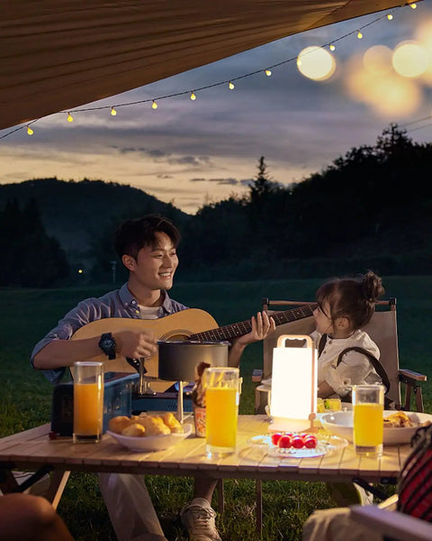 Camping scene with man playing guitar and a child, illuminated by Solar Camping Lantern With 6000mAh Rechargeable Battery Up To 80 Hours Of Light
