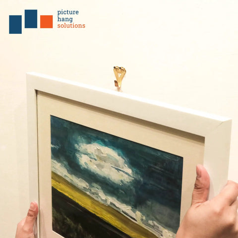 Hands hanging a framed painting using heavy duty picture hangers with nails, 50 lb wall hooks for drywall and plaster, pack of 20.