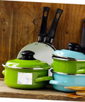 Green and blue enamel pots with lids and black-handled frying pans on wooden background
