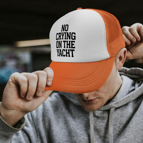 Man wearing an orange and white trucker hat with 'No Crying On The Yacht' text, adjusting the brim outdoors.