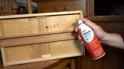 Hand holding Silicone Lubricant With Teflon Fluoro Polymer For Non Metal Materials near wooden drawer, demonstrating application.