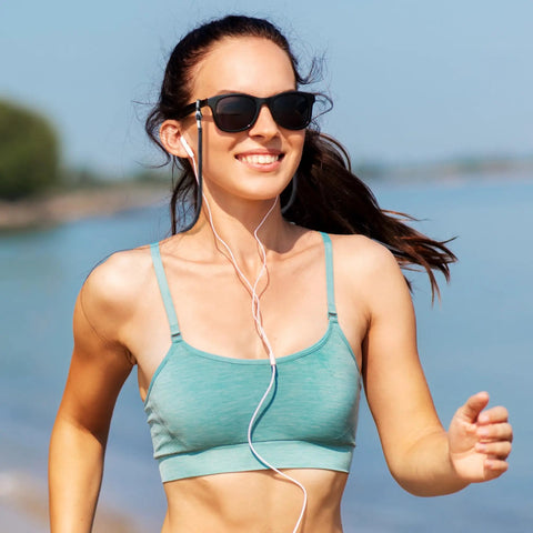 Person running on a beach wearing sunglasses and a teal sports top, listening to music with earphones.