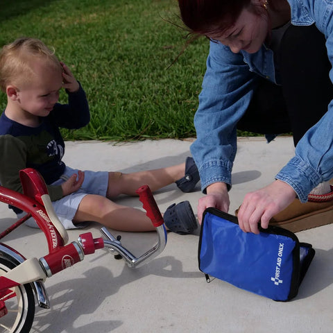 First Aid Only blue compact first-aid kit pouch on sidewalk; adult opens it beside a red tricycle and a small child.