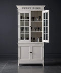 White wooden cabinet with glass doors and shelves containing glassware, set against a dark background, inscribed with 'Sweet Home'. 