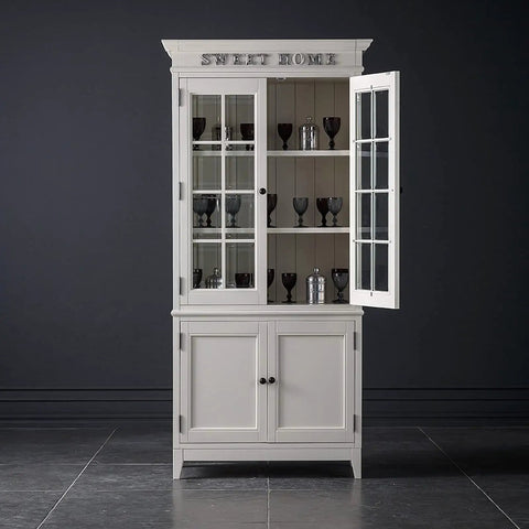 White wooden cabinet with glass doors and shelves containing glassware, set against a dark background, inscribed with 'Sweet Home'. 