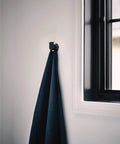 Genta LX Modern Single Robe Hook And Towel Hanger In Matte Black near window holding dark towel, space-saving bathroom accessory BH3803BL