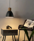 Hammered Pendant Light With Solid Wood And Metal Shade In Black Finish, above nightstand with book and chair in dim room setting.