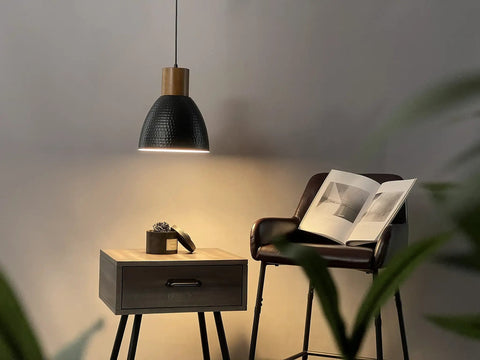 Hammered Pendant Light With Solid Wood And Metal Shade In Black Finish, above nightstand with book and chair in dim room setting.