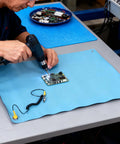 Front view of blue rubber ESD mat (26.4x20.1 in) on a workbench with grounding wire and PCB being soldered.