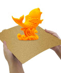 Hands holding Dual Sided Textured PEI Printing Bed For 3D Printers 235x235mm With Frosted PEI Surface with an orange 3D print.