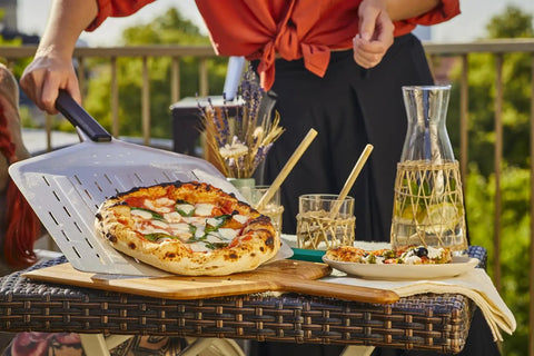 14 Inch Perforated Pizza Peel With Long Handle Aluminum Lightweight Pizza Paddle holding a pizza on an outdoor table