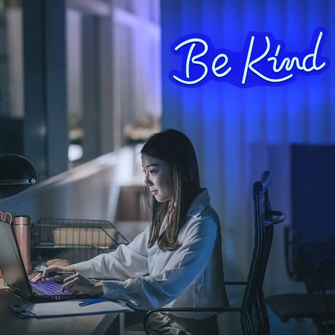 Person working at desk with blue 'Be Kind' neon sign glowing in the background.