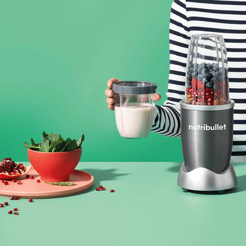 Person holding a NutriBullet 18 Ounce Short Cup with Lip Ring for Smoothies Clear Gray BPA Free Single Serve Blender Cup Without Lid Easy To Sip Mess Free Drinking Twist On Lip Ring For Convenient Single Serve Blender Accessory next to blender with ingredients.