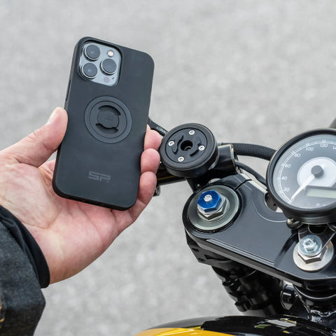 Person holding phone near SPC Plus Anti Vibration Module on motorcycle, showing mounting position and screws.