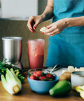 Person using a blender with 24Oz tall cup from Replacement Cup Kit for 18Oz Short, 24Oz Tall, 32Oz Colossal with Flip Top To Go Lid and Stay Fresh Resealable Lid Fits 600W and Pro 900W Blenders Dishwasher Safe, fruits and vegetables on counter.