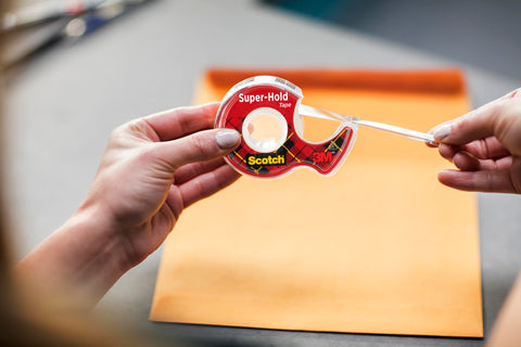 Person using Super Hold Tape With 50 Percent More Adhesive, Clear Finish, 2 Rolls, Dispenser Ready over an envelope on a table.
