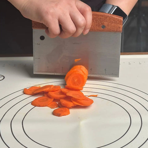 Person using Heavy Duty Bench Scraper For Flat Top Griddle, Dough Cutter And Bread Pizza Scraper to slice carrots on a board.