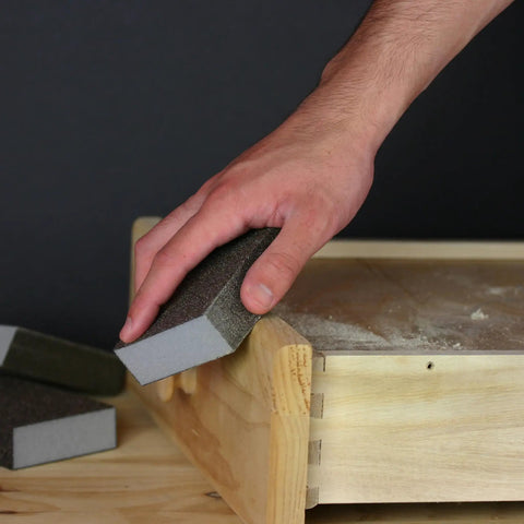 Hand using Pro Grade Sanding Sponges Pack Three By Five By One Inch Medium And Fine Grit For Contours And Flat Surfaces DIY Friendly Flexible Rinse And Reuse Aluminum Oxide Abrasive All Four Sides Ideal For Drywall Joint Finishing 2 Pack on wooden surface