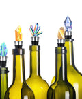 Wine bottle stoppers with metal stems and crystal gem toppers in assorted colors on green bottles (front view)