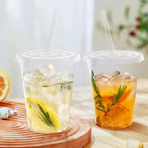 Two 100 Pack 16 Oz Clear Plastic Cups With Lids And Straws filled with iced drinks on a wooden surface.