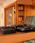 Versatile Home Audio Amplifier For Any Setup From Your Turntable To Outdoor Speakers Powers Vinyl CDs Stored Audio Files And Streaming And Even Expands Your Backyard Entertainment Black next to turntable on wooden shelf.