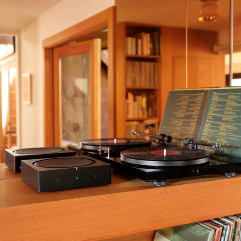Versatile Home Audio Amplifier For Any Setup From Your Turntable To Outdoor Speakers Powers Vinyl CDs Stored Audio Files And Streaming And Even Expands Your Backyard Entertainment Black next to turntable on wooden shelf.