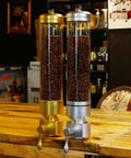 Hustle Ready Wall Mounted Coffee Bean Dispenser Keeps Your Whole Beans Crisp And Properly Locked Away Six Point Five Liter Acrylic Container With Airtight Seal, featuring gold and silver dispensers filled with coffee beans.