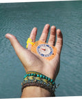 Hand holding Orienteering Compass With Luminous Pointer And Ruler For Hiking Camping And Backpacking against water backdrop.