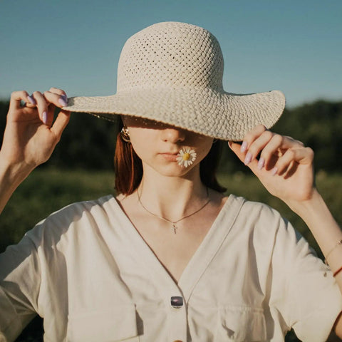 Woman in a sun hat holding the brim, with a flower in her mouth, against a serene green field backdrop.
