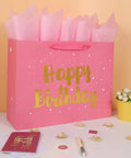 Extra Large Pink Birthday Gift Bag Set With Handles, Greeting Card, Tissue Paper And Stickers, Starry Night Design With Gold Happy Birthday Foil, Perfect For Women, Girls, And Kids, 16.5 Inch Gift Bag next to flowers and pen