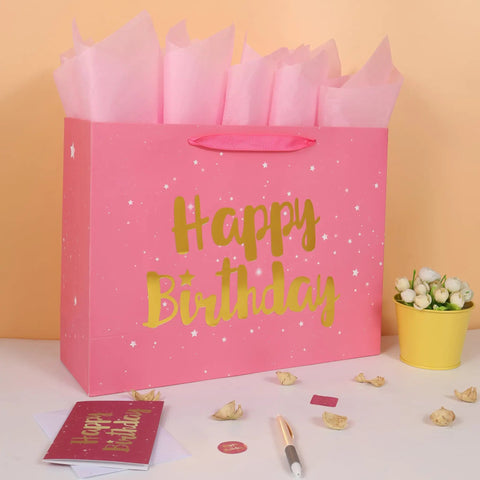Extra Large Pink Birthday Gift Bag Set With Handles, Greeting Card, Tissue Paper And Stickers, Starry Night Design With Gold Happy Birthday Foil, Perfect For Women, Girls, And Kids, 16.5 Inch Gift Bag next to flowers and pen