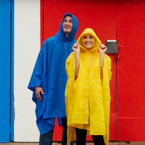 Two adults in blue and yellow ponchos smiling against a colorful wall. Lightweight Hooded Packable Poncho With Side Snaps For Adults Travel Ready Rain Gear That Packs Into Its Pouch Reusable Water Resistant Rain Shield To Keep You Dry In Any Forecast.