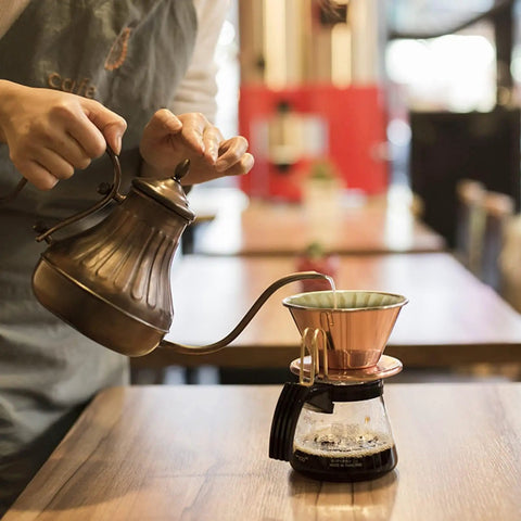 Person pouring hot water over coffee filter in a cafe setting, illustrating use of 100 Pack 1 To 4 Cup Disposable Basket Coffee Filters For A Smooth Brew Efficient And Convenient 1 Cup Size Eco Friendly Biodegradable Natural Pulp With Wavy Edges And Flat Bottom For Even Filtration
