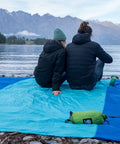Two people sitting on a Sandproof Beach Blanket With 6 Stakes And 4 Corner Pockets Large 7 By 7 Feet 210 Cm Square by a lake.