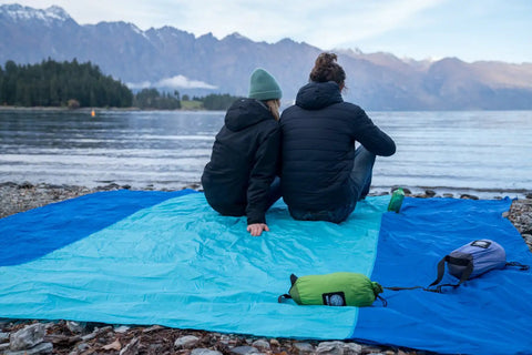 Two people sitting on a Sandproof Beach Blanket With 6 Stakes And 4 Corner Pockets Large 7 By 7 Feet 210 Cm Square by a lake.