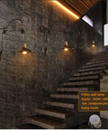 Dope Vintage Wall Sconce Featuring Classic Wrought Iron Build With Handsome Pulley Adjustment For Slick Loft Rooms And Chill Bar Decor That Seriously Slays And Looks Absolutely Lit on a stone wall next to stairs.