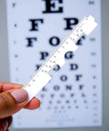 Two Piece White Pupil Distance Ruler For Eye Care And Prescription Eyeglasses PD Meter With Monocular And Binocular Measurement Options And Easy Read Scale, held in front of eye chart