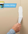 Hand using putty knife with Alex Flex Flexible Spackling White 16 Oz Won’t Sink Shrink Or Crack Sandable And Paintable Formula on wall