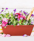 Red 17-inch rectangular plastic window box planter with purple and pink flowers and a gardening tool visible on top.