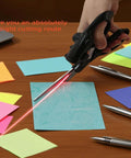 Laser-guided fabric scissors with stainless blades, handheld; cutting blue sticky note with red laser line visible.