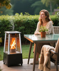 Outdoor patio heater beside a glass-top table with a seated woman; metal freestanding unit with glass enclosure.