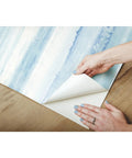 Hands peeling Tan Watercolor Stripe Peel And Stick Wallpaper Roll 20.5 Inch Wide By 16.5 Foot Long for easy DIY installation on wood.